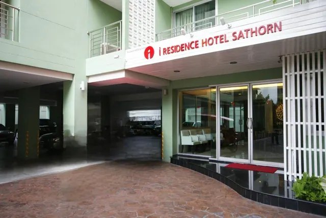 Exterior of the i Residence Hotel Sathorn showing red signage, a glass entrance with seating visible inside, and covered parking beneath the building.