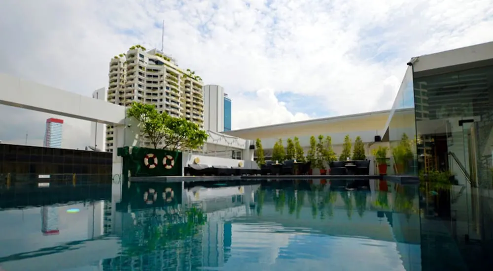 A rooftop swimming pool with reflections on the water, lounge seating and potted plants, with high-rise towers visible behind.