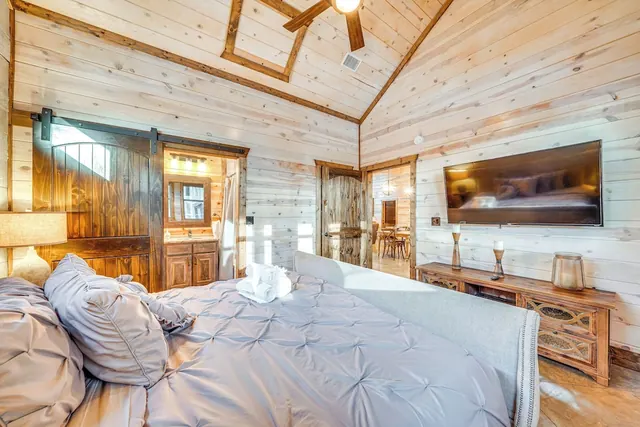 A bedroom with a made bed, wood plank walls and ceiling, a wall-mounted television, a ceiling fan and a wooden dresser, with a barn door to the bathroom on the left.