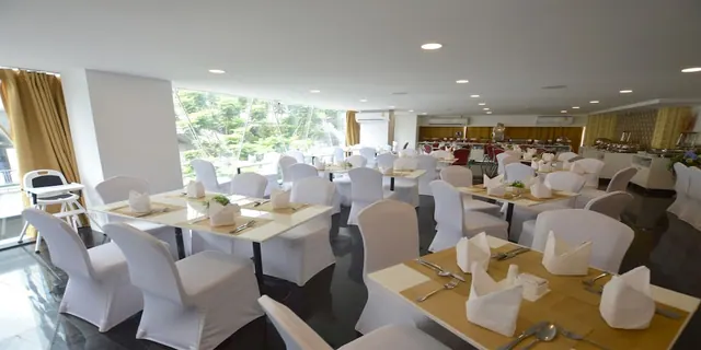 A dining room with white-covered chairs and set tables, a buffet counter at the back, and large windows looking onto trees.