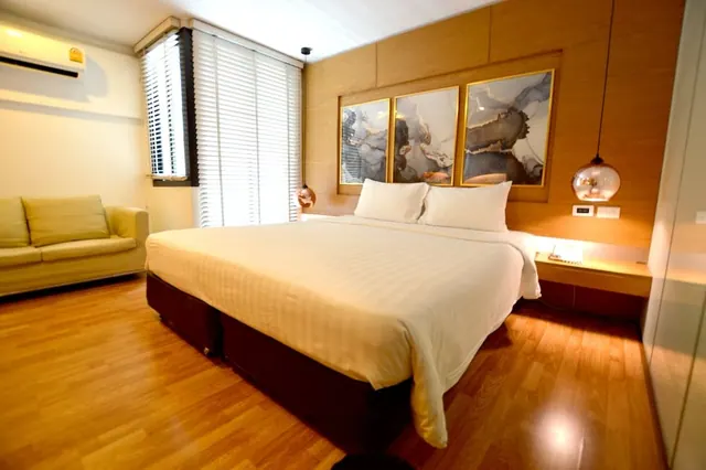 A bedroom with a large king bed dressed in white linen, a beige sofa on the left, a wall-mounted air conditioner and three framed abstract artworks above the headboard.