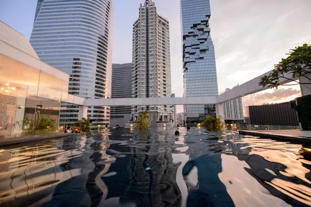 A rooftop pool at dusk with a few people swimming, with tall glass towers rising behind it.