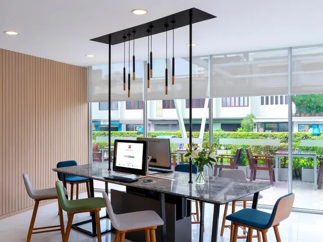 A communal work table with a monitor, keyboard and coloured chairs sits beneath black pendant lights in a bright area with floor-to-ceiling windows onto a street.