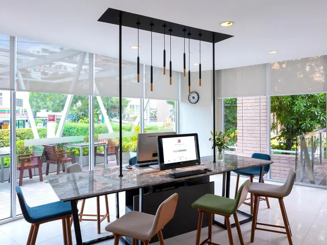 A lobby web corner with a marble-top counter, two computer monitors, high stools and pendant lights, with large windows looking onto trees and an outdoor seating area.