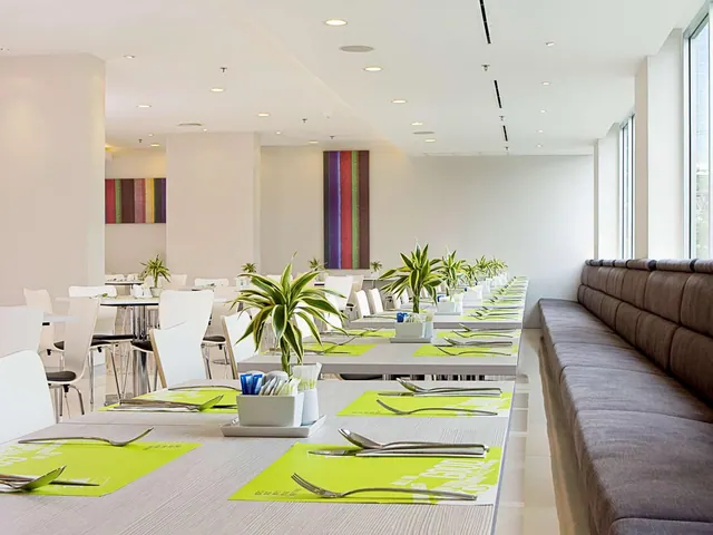Long rows of dining tables with lime green placemats and white chairs run through a bright dining room, with a brown upholstered banquette along the right wall.