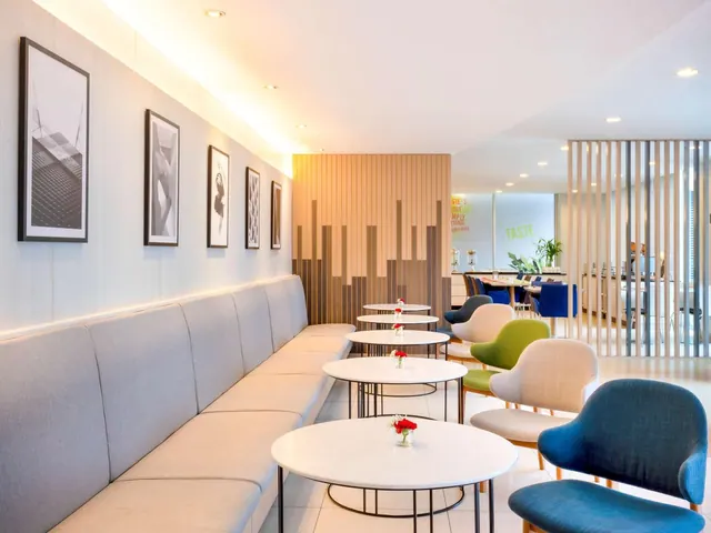 A lounge seating area with a long grey banquette, round white tables with small flower vases, coloured armchairs and framed black and white prints on the wall.