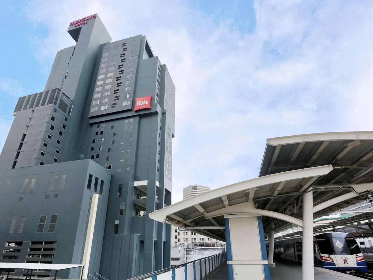 Low-angle view of a tall grey hotel tower with red signage beside an elevated Skytrain platform and a passing train.