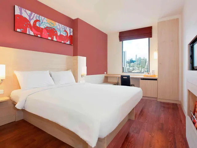 A bedroom with a king bed, a work desk, a wardrobe and a city skyline visible through the window.