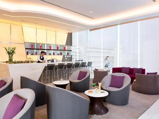 A lounge with a bar counter where a bartender stands, surrounded by grey armchairs with purple cushions, small round tables and a row of tall windows.
