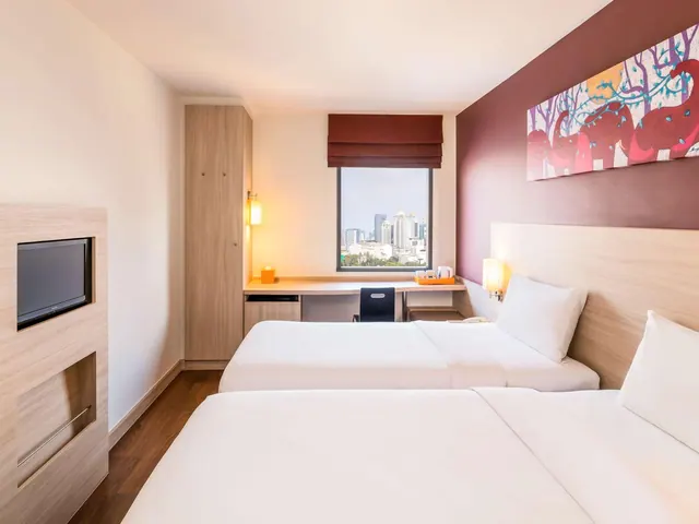 A hotel bedroom with two twin beds, a desk and chair, a wall-mounted television, and a window showing a city skyline.