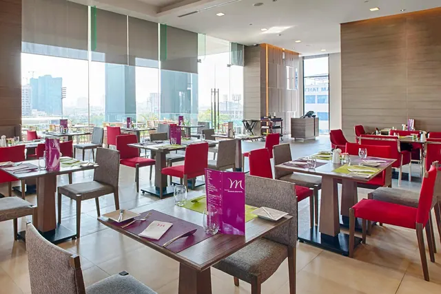 A restaurant dining room with red and grey chairs at wooden tables set with place settings, and floor-to-ceiling windows looking out over city buildings.