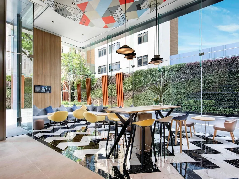 A lobby seating area with a grey sofa, yellow chairs and high tables on a black and white marble floor, with a green wall visible through the glass.