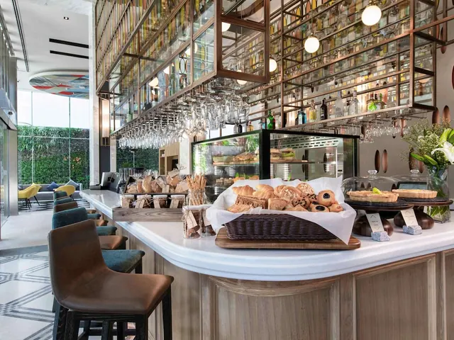 A bar counter with a pastry basket and display case sits beneath a wall of glassware and bottles, with lounge armchairs and a green wall visible through the windows beyond.