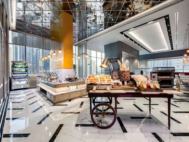A restaurant breakfast area with a marble floor, a bread display cart, a buffet counter and large windows showing high-rise buildings.