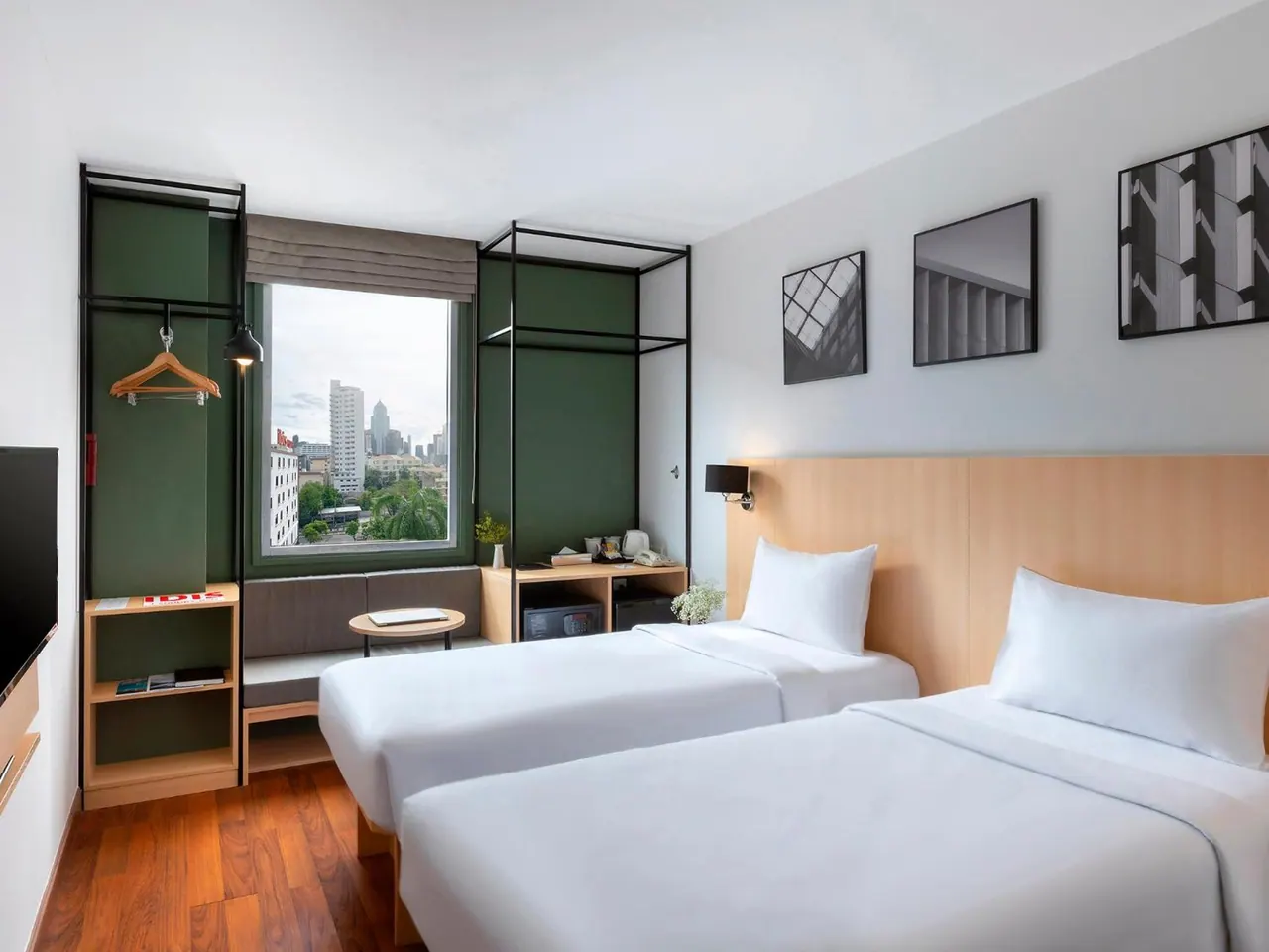 A bedroom with two single beds, a green accent wall, a desk, a wall-mounted television and a window looking onto city buildings.