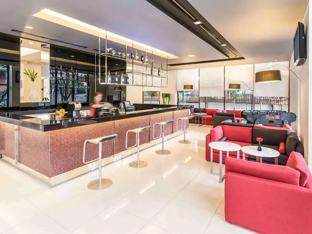 A bar counter with a mosaic-tiled front and stools sits beside red sofas and a foosball table in a lounge with large windows.