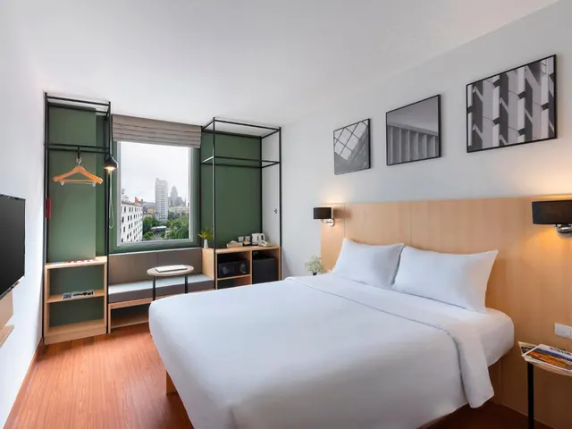 A modern hotel bedroom with a double bed, a built-in bench by a window with a view of city buildings, an open clothes rail, and a tea station with a kettle.