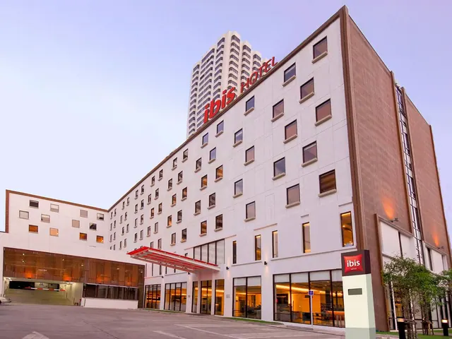 The exterior of an ibis hotel building with a red entrance canopy, illuminated windows and a taller tower behind it at dusk.