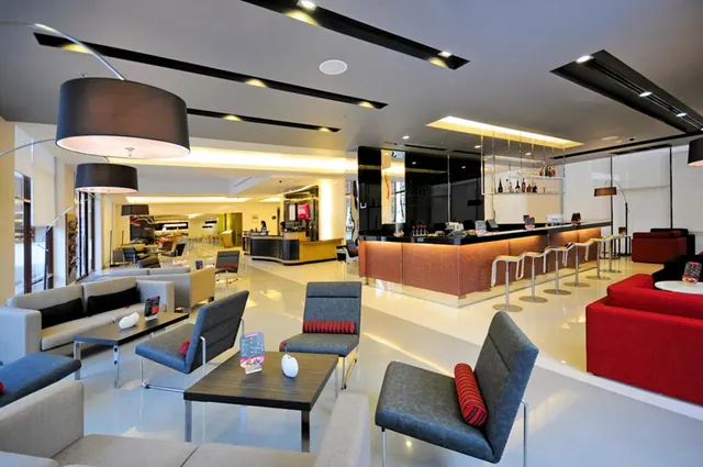A hotel lounge with grey armchairs, red sofas and a bar counter with stools beneath large pendant lamps.