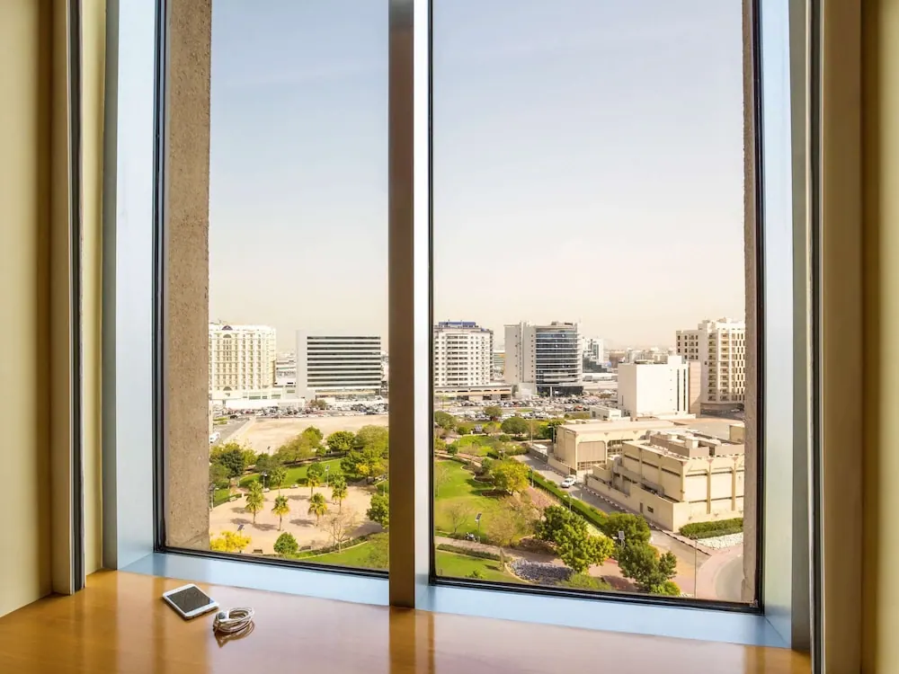 Tall windows look out over a park and a city of mid-rise and high-rise buildings under a pale hazy sky, with a phone and earphones on a wooden ledge in front.