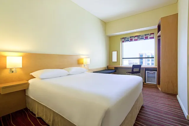 A large bed with white bedding and two wall lamps sits beside a wooden headboard, with a desk, chair and minibar near a window looking out onto nearby buildings.