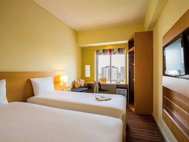 A bedroom with two single beds with white bedding, a desk and chair by a window overlooking city buildings, and a wall-mounted television.