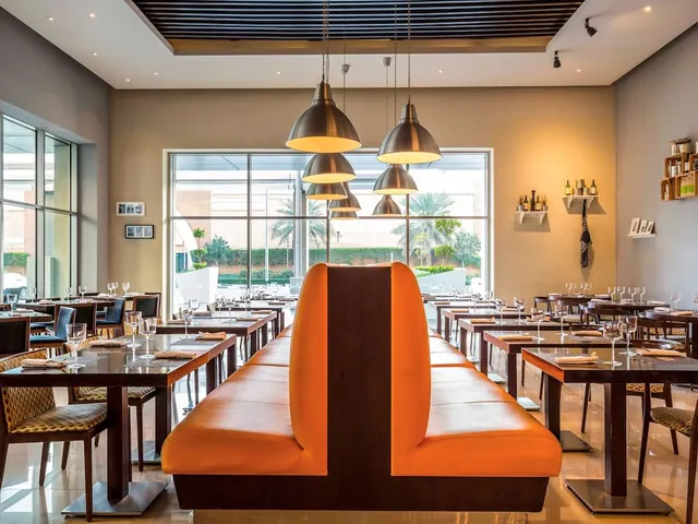A restaurant dining room with tables set for dining, an orange leather banquette in the foreground, pendant lamps overhead, and large windows looking out onto palm trees and hedges.