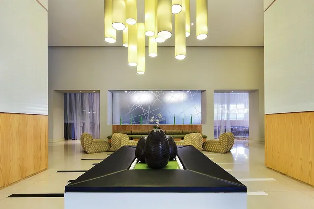 A hotel lobby with suspended yellow cylindrical lights, a black central island holding a large dark vase with white flowers, and upholstered lounge seating along the back wall.