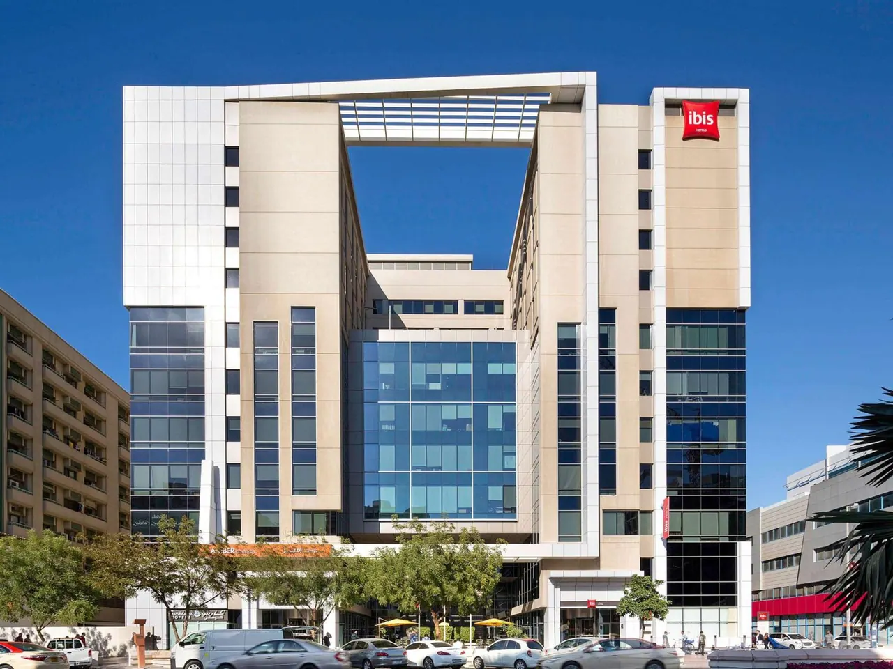 Exterior view of a multi-storey ibis hotel building with a red ibis sign on its facade, cars on the street and trees in front, under a clear blue sky.