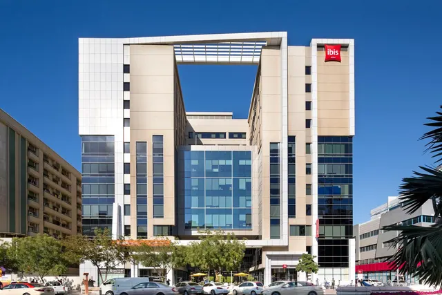Front exterior of an ibis hotel building under a clear blue sky, with cars parked on the street and trees in front.