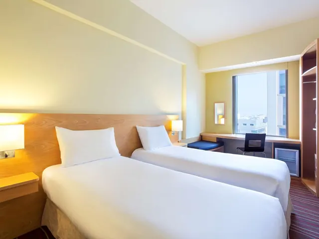 A twin bedroom with two single beds, a desk by a window looking out onto buildings, and a wardrobe and minibar on the right.