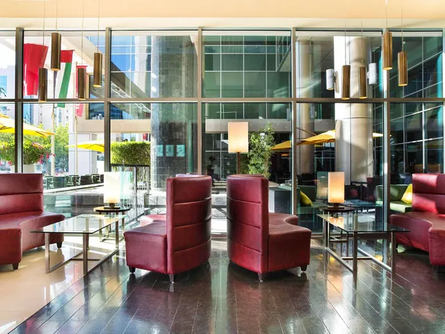 A lobby lounge with red leather armchairs and glass coffee tables sits in front of floor-to-ceiling windows that look out onto an outdoor area with yellow umbrellas and buildings.