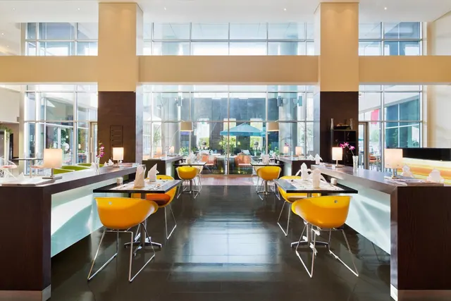 A restaurant dining room with yellow chairs, set tables with table lamps and folded napkins, and floor-to-ceiling windows along the back wall.