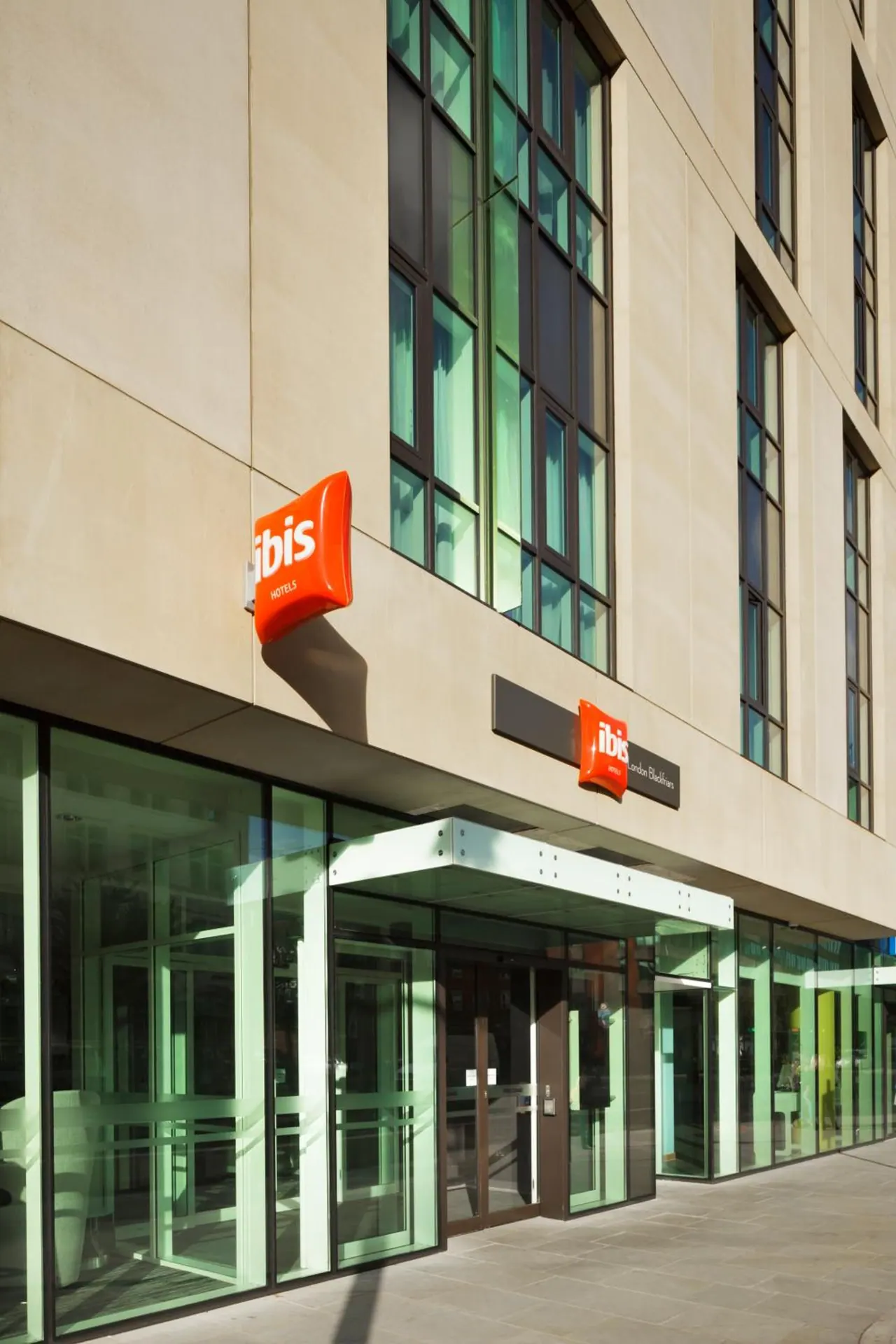 The exterior of an ibis hotel shows a glass entrance canopy, orange ibis signs on a stone facade and a paved pavement in front.