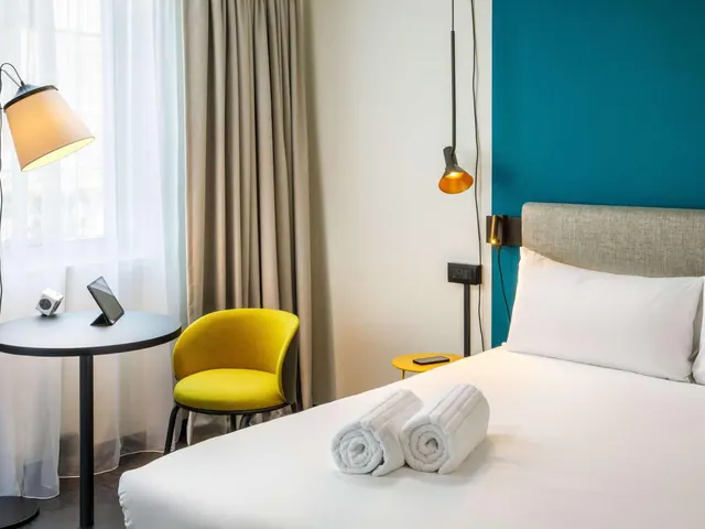 A made double bed with two rolled white towels on it, a yellow armchair and a round table by a sheer-curtained window, and a teal accent wall behind the headboard.