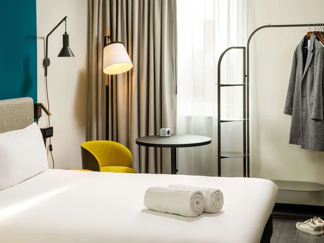 A hotel bedroom with a made bed, two rolled white towels on the bed, a yellow armchair, a round side table, a floor lamp and a coat hanging on a rail.