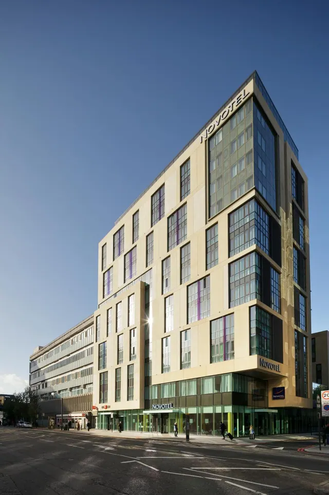 Exterior view of a tall cream-coloured Novotel hotel building with glass lower floors under a clear blue sky, with a road and a few pedestrians at street level.