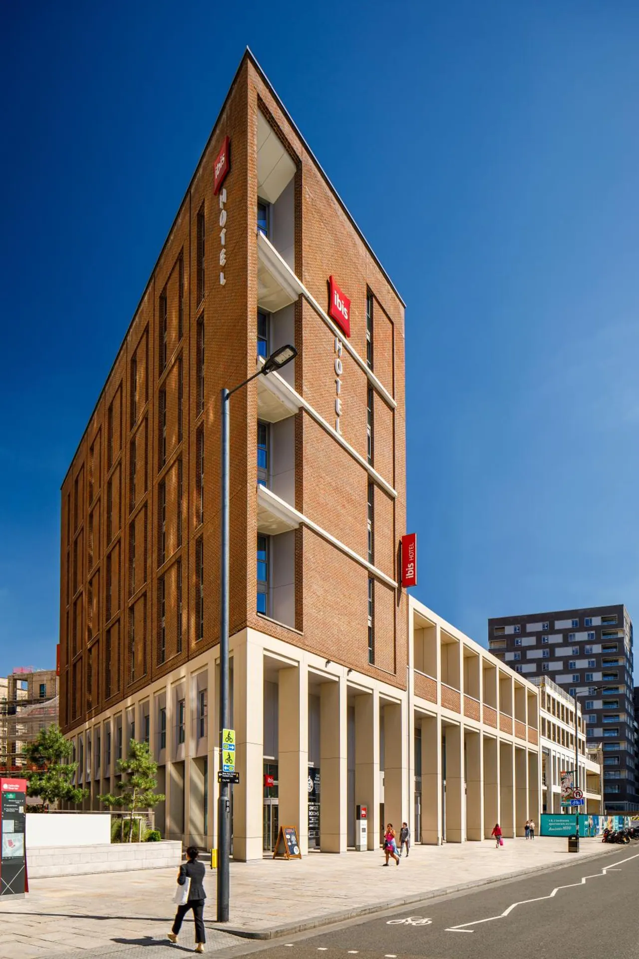 Exterior view of a tall brick ibis hotel building with a tapered upper section and red signage, a street lamp and a few pedestrians on the pavement under a clear blue sky.