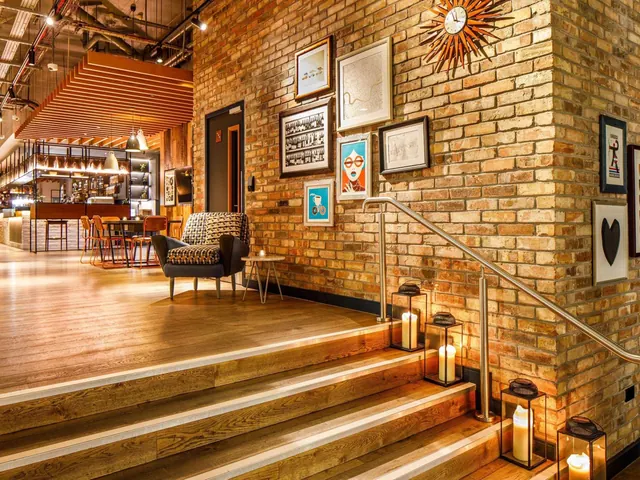 A lobby seating area with an armchair, framed prints on a brick wall, and a wooden staircase lined with lit candle lanterns, with a bar and dining tables visible in the background.
