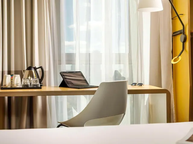 A wooden desk with a tablet, sunglasses, and a kettle and cups on a tray sits by a sheer-curtained window, with a grey chair and the edge of a white bed in the foreground.