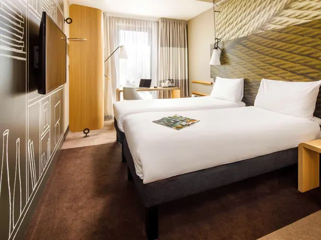 A hotel bedroom with two single beds with white bedding, a wall-mounted television, and a desk with a laptop by the window.