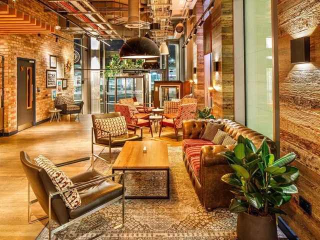 A lobby lounge with a brown leather Chesterfield sofa and leather armchairs around a wooden coffee table, under pendant lights, with brick walls and potted plants.