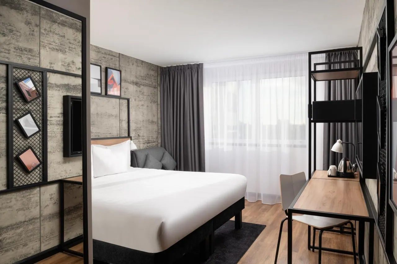 A double bed with white linen sits beside a grey armchair, a wooden desk with a chair, and a kettle on the desk, with a concrete-effect feature wall and sheer curtains on the window.