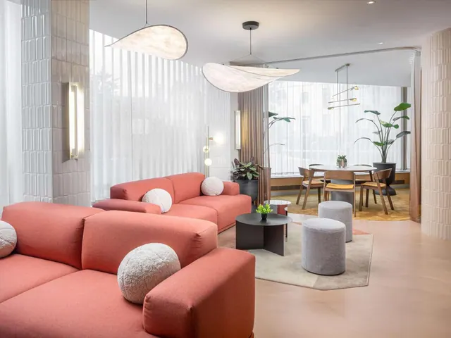 A lounge seating area with coral sofas, grey stools, pendant lights and a round dining table with chairs in the background.