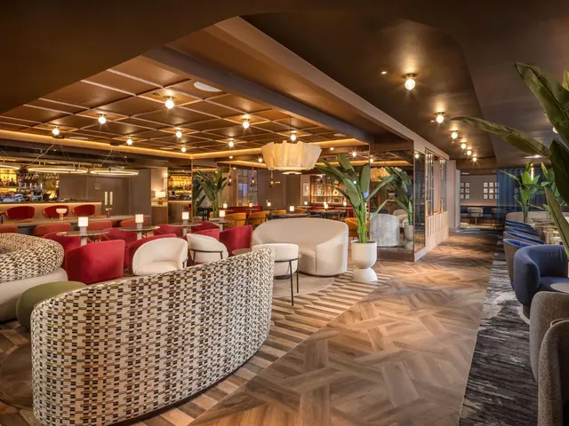 A lounge with a bar on the left, curved upholstered seating, red chairs, potted plants and pendant lights over a parquet floor.