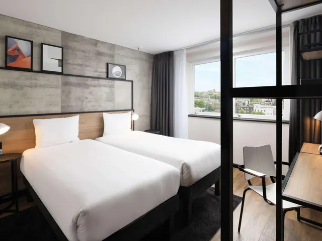 A bedroom with two single beds with white bedding, a wooden desk and chair, and a window looking out over city rooftops.