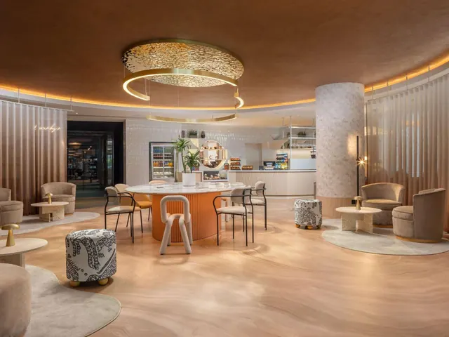 A lobby lounge with a round marble-topped table, upholstered armchairs, patterned stools and a pastry counter in the background.