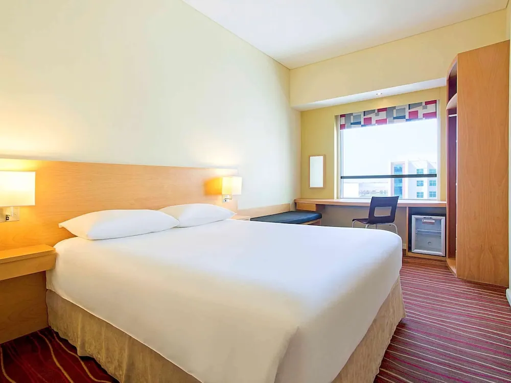 A bedroom with a double bed, wooden headboard and bedside lamps, a desk and chair by the window, and a wardrobe and minibar fridge.