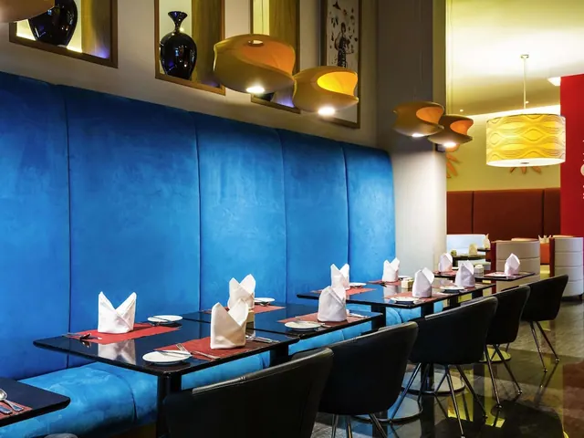 A restaurant dining room with a long blue upholstered banquette, black tables set with folded white napkins and red placemats, black chairs, and pendant lights.