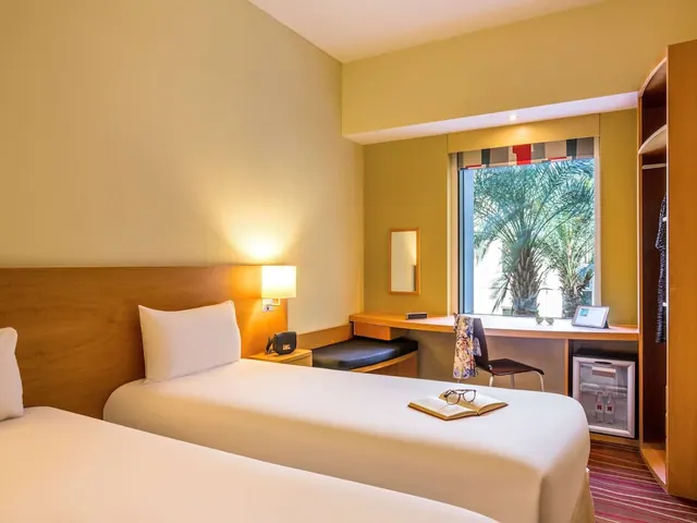A twin-bedded hotel room with a desk under a window looking out onto palm trees, with a wardrobe and a minibar.